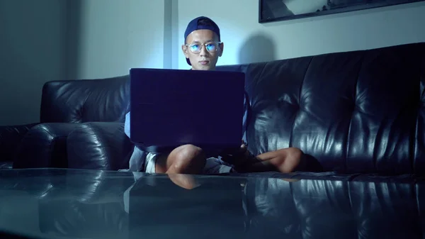 Joven hombre asiático guapo en gafas con reflejos usando su computadora portátil, sentado en la noche en la habitación. En la oscuridad —  Fotos de Stock