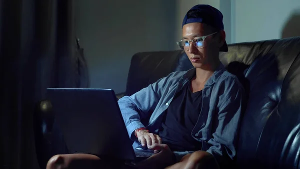 Молодой красивый азиат в очках с размышлениями за ноутбуком, сидящий вечером в комнате. В темноте — стоковое фото
