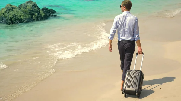 Ein junger Mann in Hose und klassischem Hemd mit Koffer spaziert vor der Kulisse des türkisfarbenen Meeres am Strand entlang. freiberufliches Konzept, Ruhe — Stockfoto