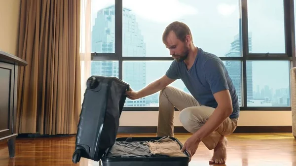 Homem bonito embala uma mala em um quarto com uma janela panorâmica com vista para os arranha-céus — Fotografia de Stock