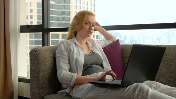 Una mujer elegante está acostada en un sofá junto a la ventana panorámica con vistas a los rascacielos y el uso de su ordenador portátil —  Fotos de Stock