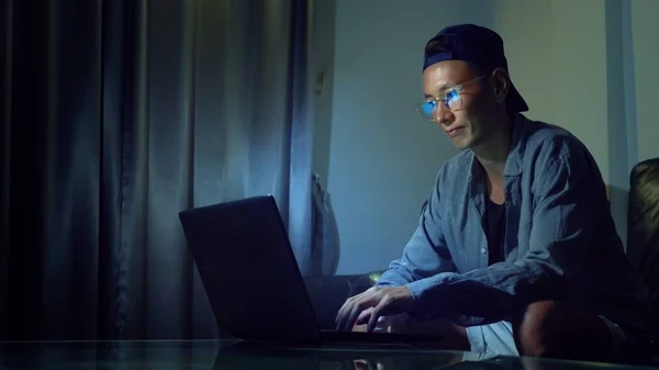 Jovem asiático bonito em óculos com reflexos usando seu laptop, sentado à noite na sala. No escuro — Fotografia de Stock