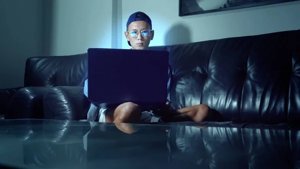 Joven hombre asiático guapo en gafas con reflejos usando su computadora portátil, sentado en la noche en la habitación. En la oscuridad — Foto de Stock