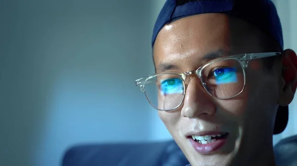 Giovane bell'uomo asiatico in occhiali con riflessi utilizzando il suo computer portatile, seduto la sera nella stanza. Al buio. — Foto Stock