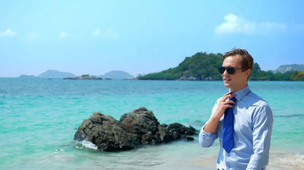 Homem de negócios bonito em óculos de sol caminhou ao longo de uma praia tropical, tirando sua gravata — Fotografia de Stock