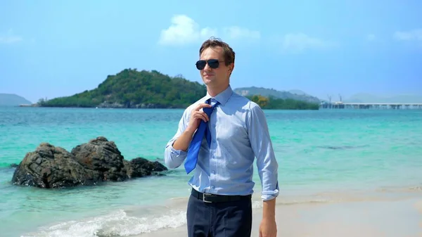 Homem de negócios bonito em óculos de sol caminhou ao longo de uma praia tropical, tirando sua gravata — Fotografia de Stock