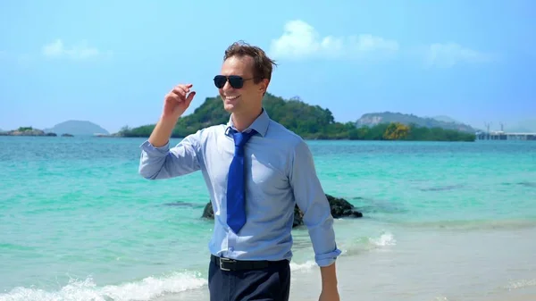 Homem de negócios bonito em óculos de sol caminhou ao longo de uma praia tropical, tirando sua gravata — Fotografia de Stock