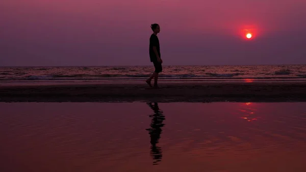 Silueta. solitario asiático joven caminando pacíficamente a lo largo de una playa desierta al atardecer. paisaje marino — Foto de Stock