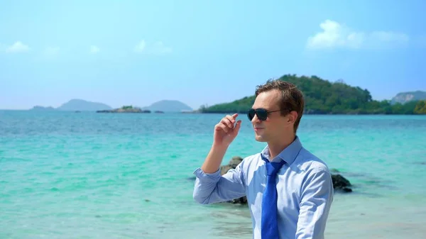 Επιχειρηματία σε ένα πουκάμισο και γραβάτα, σε γυαλιά ηλίου πηγαίνει σε μια λευκή αμμώδη παραλία. Freelancing έννοια, διακοπές — Φωτογραφία Αρχείου