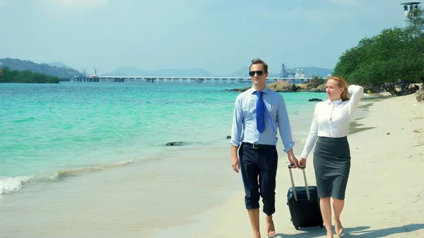 Homem de negócios e mulher de negócios com uma mala andando ao longo da praia de areia branca na ilha — Fotografia de Stock