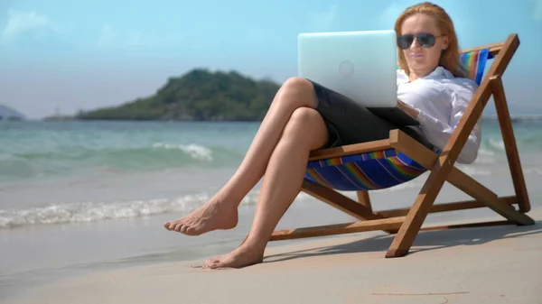 Empresaria que trabaja en un ordenador portátil mientras está sentada en una tumbona junto al mar en una playa de arena blanca. concepto freelance o workaholism . —  Fotos de Stock