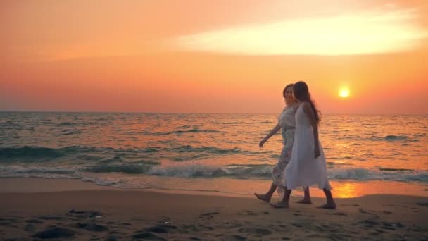 Moeder en dochter in witte jurken lopen blootsvoets op een zandstrand, hand in hand tegen de achtergrond van een schitterende zonsondergang — Stockvideo
