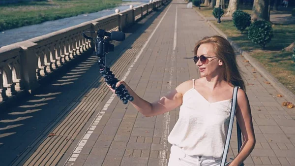 Hermosa bloguera feliz mujer con una cámara, conduce su reportaje paseando por el terraplén de la ciudad turística. el viento desarrolla su cabello, la luz del sol brilla a través de las ramas de los árboles . — Foto de Stock