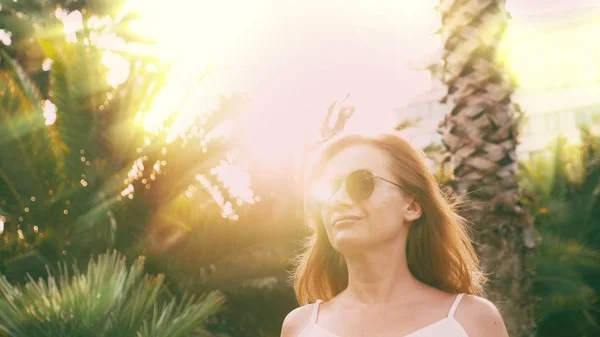 Bella donna bionda elegante in occhiali da sole, passeggiando lungo un sentiero di palme. Il palmo si riflette nei bicchieri . — Foto Stock