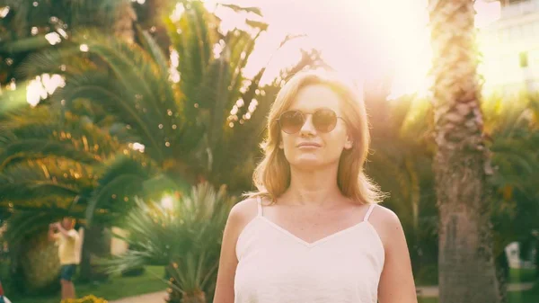 Mooie stijlvolle blonde vrouw in zonnebril, wandelen langs een palm tree pad. De palm wordt weerspiegeld in de glazen. — Stockfoto