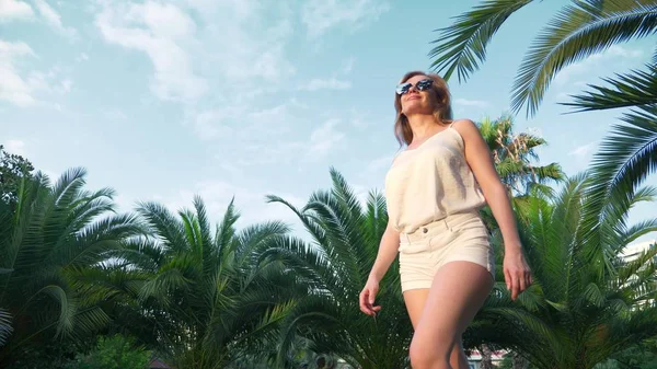 Kaunis tyylikäs vaalea nainen aurinkolaseissa, kävely palmupolkua pitkin. Kämmen heijastuu laseista . — kuvapankkivalokuva