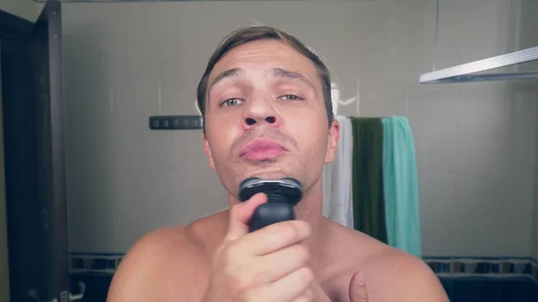 Yakışıklı bir genç adam banyoda ayna önünde duran bir elektrikli tıraş makinesi ile sakalını tıraş. — Stok fotoğraf