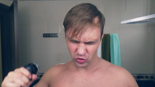 Un joven guapo se afeita la barba con una afeitadora eléctrica, de pie en el baño frente al espejo . — Foto de Stock