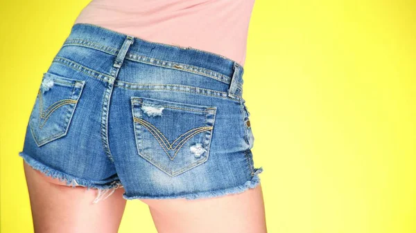 Chica en pantalones cortos pantalones cortos azules. Vista de cerca desde atrás. Estilo fresco de verano. fondo amarillo de color —  Fotos de Stock