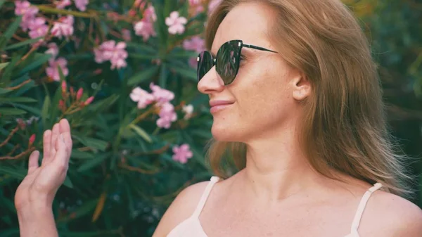 Beautiful stylish blond woman in sunglasses walking in the park with flowering shrubs. oleander flowers are reflected in the glasses. — Stock Image Beautiful stylish blond woman in sunglasses walking in the park with flowering shrubs. oleander flowers are reflected in the glasses. Stock Photo