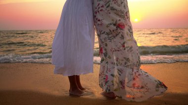 mother and daughter in white dresses walk barefoot on a sandy beach, holding hands against the backdrop of a magnificent sunset
