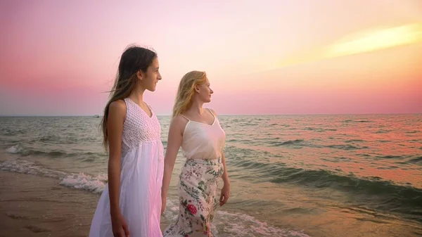 Madre e hija en vestidos blancos caminan descalzas en una playa de arena, tomados de la mano contra el telón de fondo de una magnífica puesta de sol — Foto de Stock