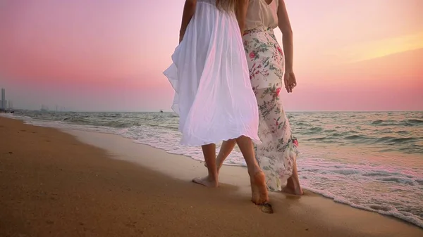 Madre e hija en vestidos blancos caminan descalzas en una playa de arena, tomados de la mano contra el telón de fondo de una magnífica puesta de sol — Foto de Stock