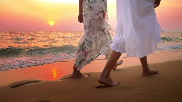 De cerca. pies desnudos en la playa. dos mujeres jóvenes en vestido blanco caminando por la playa al atardecer — Foto de Stock