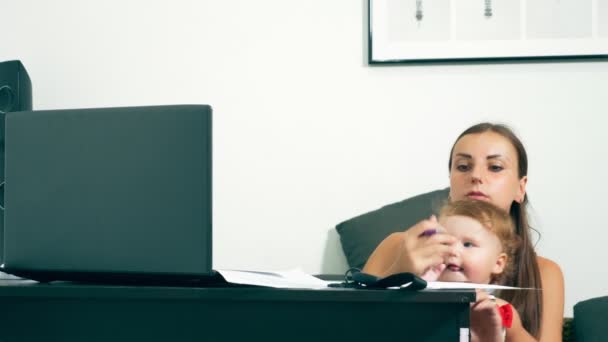 Concepto de trabajo independiente femenino. La maternidad moderna. Madre trabajadora con un hijo en la mesa. Mujer ocupada trabajando en un portátil con un bebé en sus brazos . — Vídeo de stock