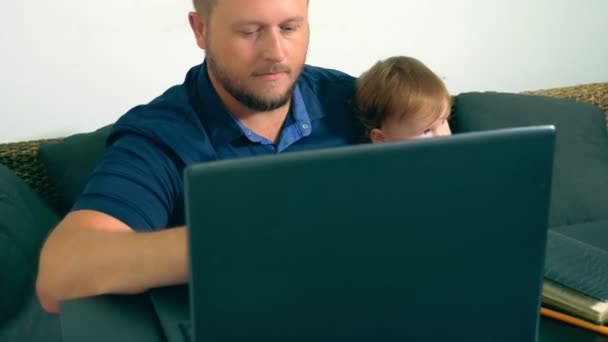 フリーランサーの仕事のコンセプトです。彼の腕の中で赤ん坊を保持している、ノート パソコンと一緒に家で働く幸せお父さん. — ストック動画
