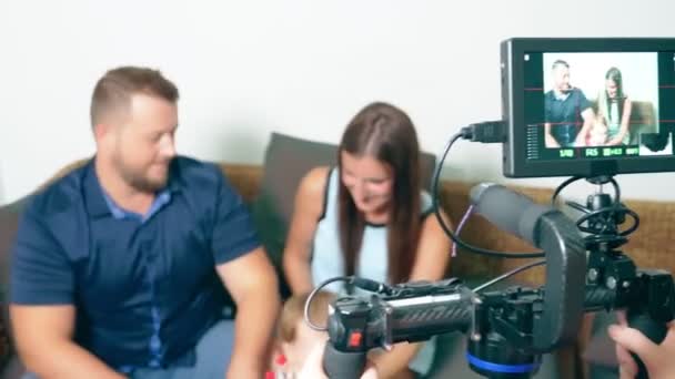 Proceso de grabación de vídeo. Un operador con un steadicam dispara una historia sobre una familia, mamá y papá con una niña pequeña en un acogedor sofá en casa, que charlan entre sí — Vídeo de stock