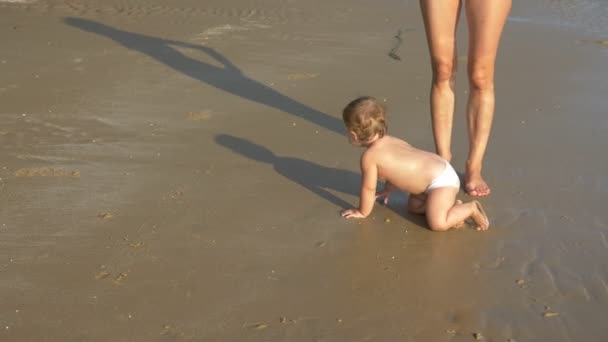 Loira bebê rastejando na praia de areia, mamãe feliz olha para ela — Vídeo de Stock