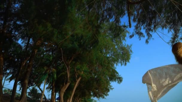 Una chica rubia feliz en un vestido blanco y capa de encaje se balancea en un columpio de cuerda, en la playa junto al mar . — Vídeo de stock