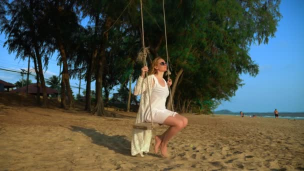 Una chica rubia feliz en un vestido blanco y capa de encaje se balancea en un columpio de cuerda, en la playa junto al mar . — Vídeos de Stock