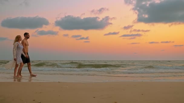 Casal romântico, homem e mulher caminham ao longo da praia ao pôr do sol, de mãos dadas . — Vídeo de Stock