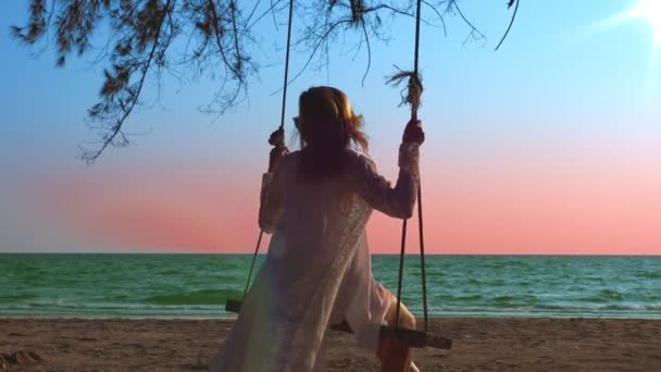 Una ragazza bionda felice con un vestito bianco e un mantello di pizzo sta oscillando su un'altalena di corda, sulla spiaggia vicino al mare . — Video Stock