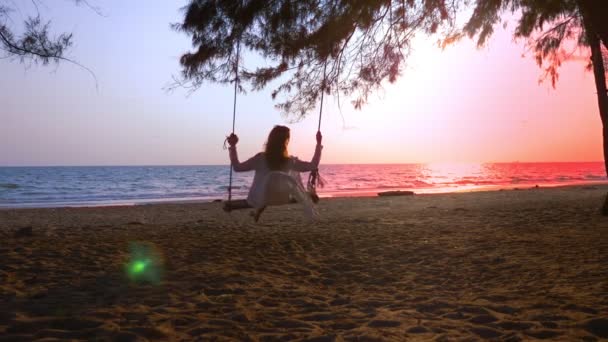 Ein glückliches blondes Mädchen in weißem Kleid und Spitzenumhang schwingt auf einer Seilschaukel am Strand am Meer. — Stockvideo