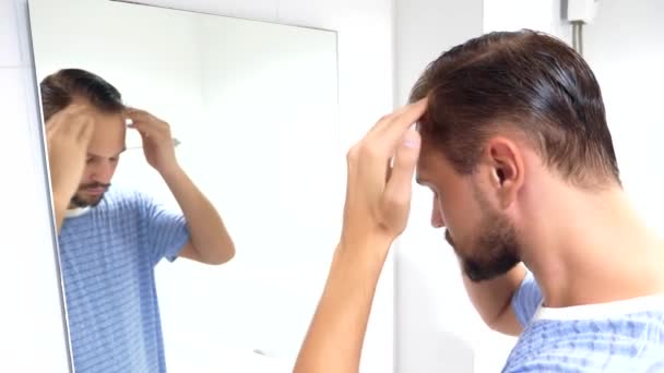 A young man with the problem of hair loss, looking in the mirror indoors, massages the skin of the head — Stock Video