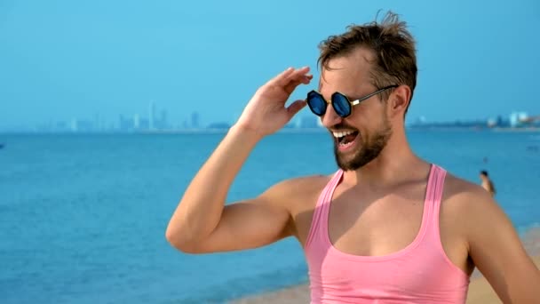 Gros plan, beau gosse ludique en t-shirt rose sur une plage tropicale. il regarde la caméra, se réjouit et fait des grimaces drôles — Video