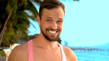 Yakın çekim, tropikal bir plajda pembe tişört oynak yakışıklı adam. o da kameraya baktı, sevinir ve komik yüz yapıyor