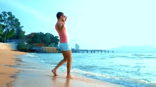 Un bel ragazzo giocherellone con una t-shirt rosa e pantaloncini blu corre gioiosamente in mare. mostro sul mare — Video Stock