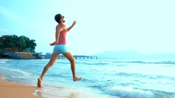 Un chico guapo juguetón con una camiseta rosa y pantalones cortos azules corre alegremente hacia el mar. monstruo en el mar — Vídeos de Stock