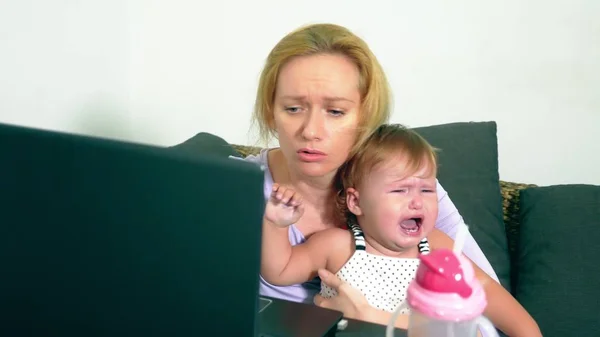 Annoyed businesswoman holding a crying baby in her arms while working on a laptop. Career mom concept, problems associated with work at home