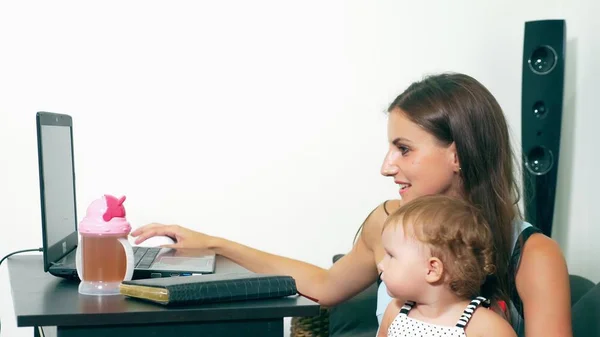 Female freelance job concept. Modern motherhood. Working mother with a child at the table. Busy woman working on a laptop with a baby in her arms.