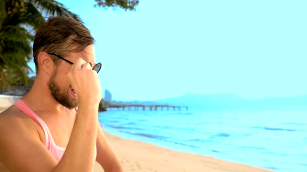 Close-up, speelse knappe jongen in roze t-shirt op een tropisch strand. Hij kijkt naar de camera, verheugt zich en maakt grappige gezichten — Stockvideo