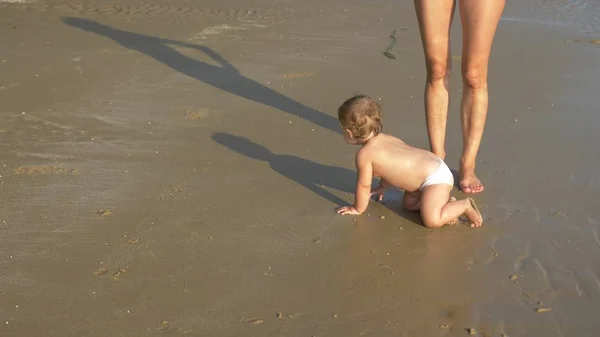 Блондинка Дитина плаче на піщаному пляжі, мама щасливо дивиться на неї — стокове фото