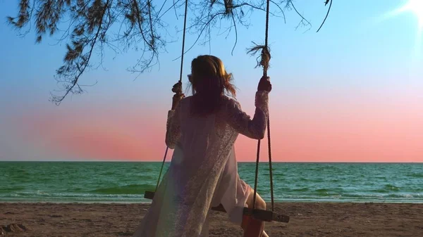 Uma menina loira feliz em um vestido branco e capa de renda está balançando em um balanço de corda, na praia junto ao mar . — Fotografia de Stock