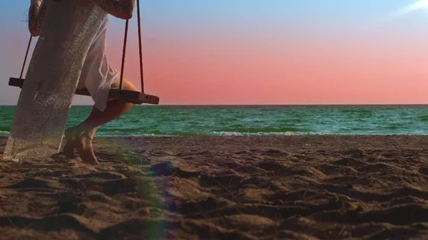 Une fille blonde heureuse en robe blanche et cape en dentelle se balance sur une balançoire à corde, sur la plage au bord de la mer . — Photo