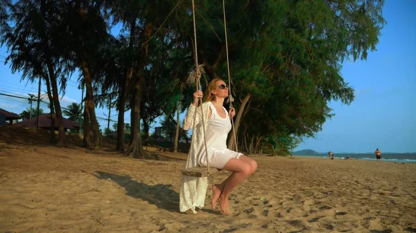 Seorang gadis pirang yang bahagia dengan gaun putih dan jubah renda berayun di ayunan tali, di pantai dekat laut . — Stok Foto