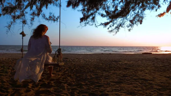 Una chica rubia feliz en un vestido blanco y capa de encaje se balancea en un columpio de cuerda, en la playa junto al mar . — Foto de Stock